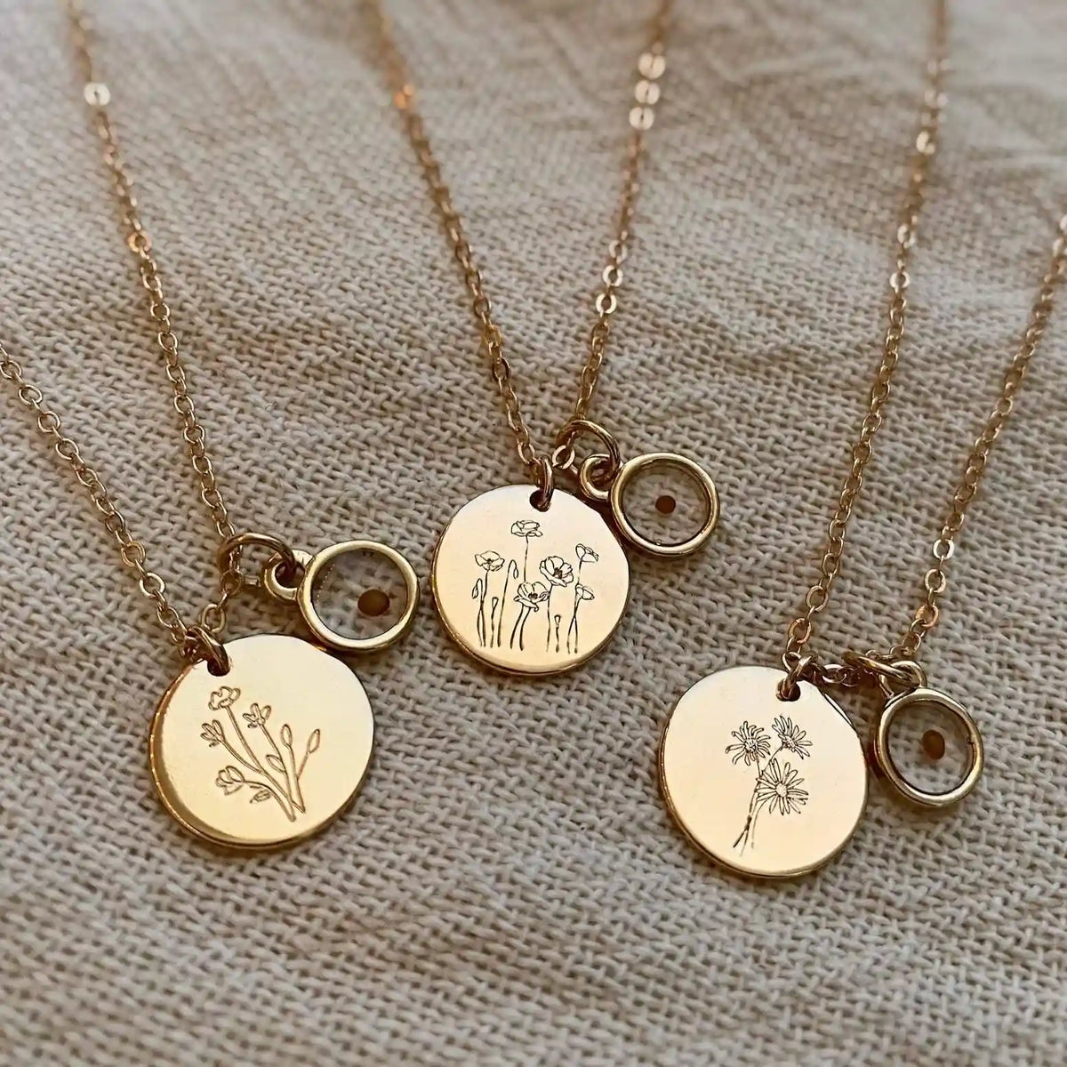 Three gold necklaces with circular pendants featuring floral designs on a textured fabric background.