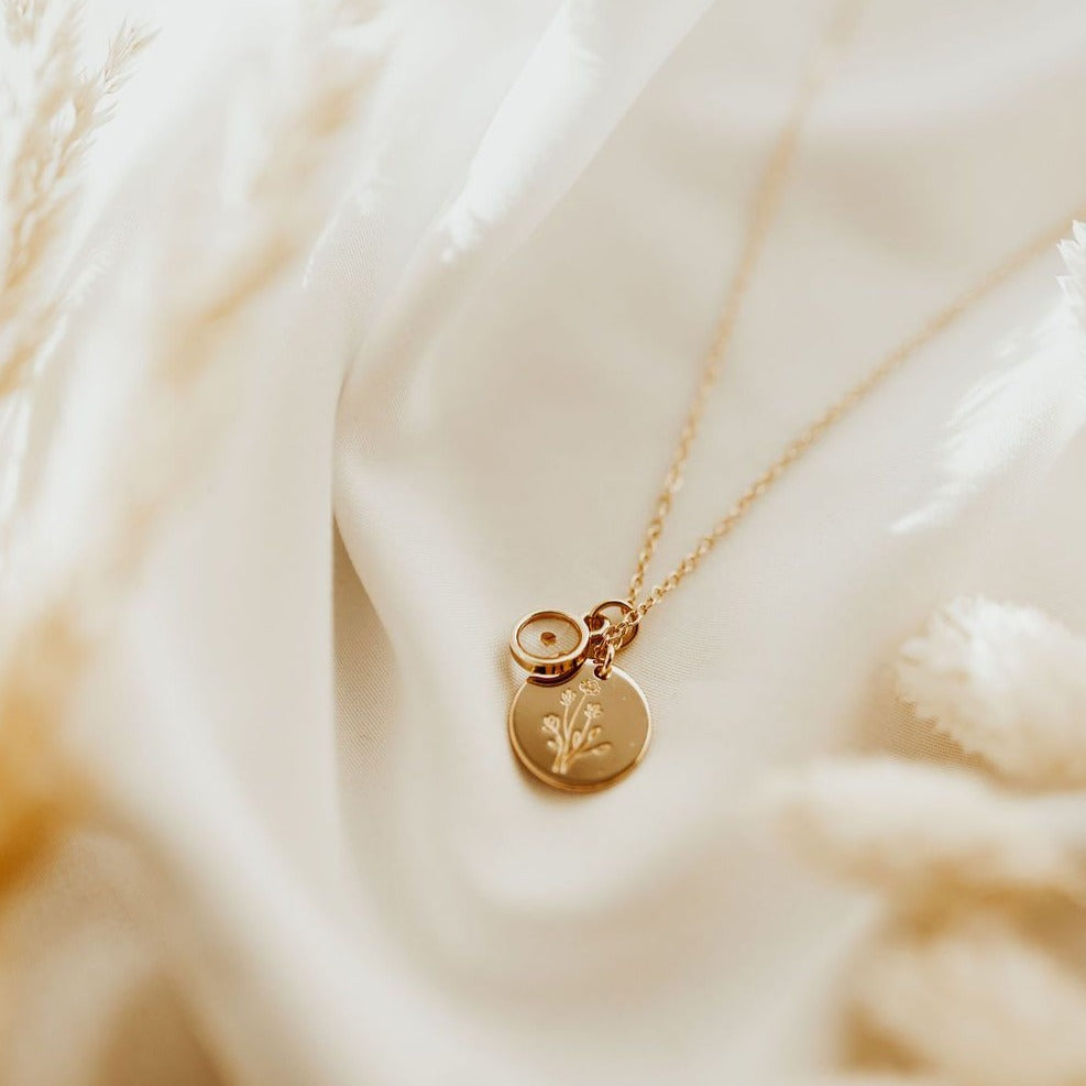 A gold-plated necklace with a small wildflower charm and a mustard seed pendant on achain laid out on a soft, textured surface.